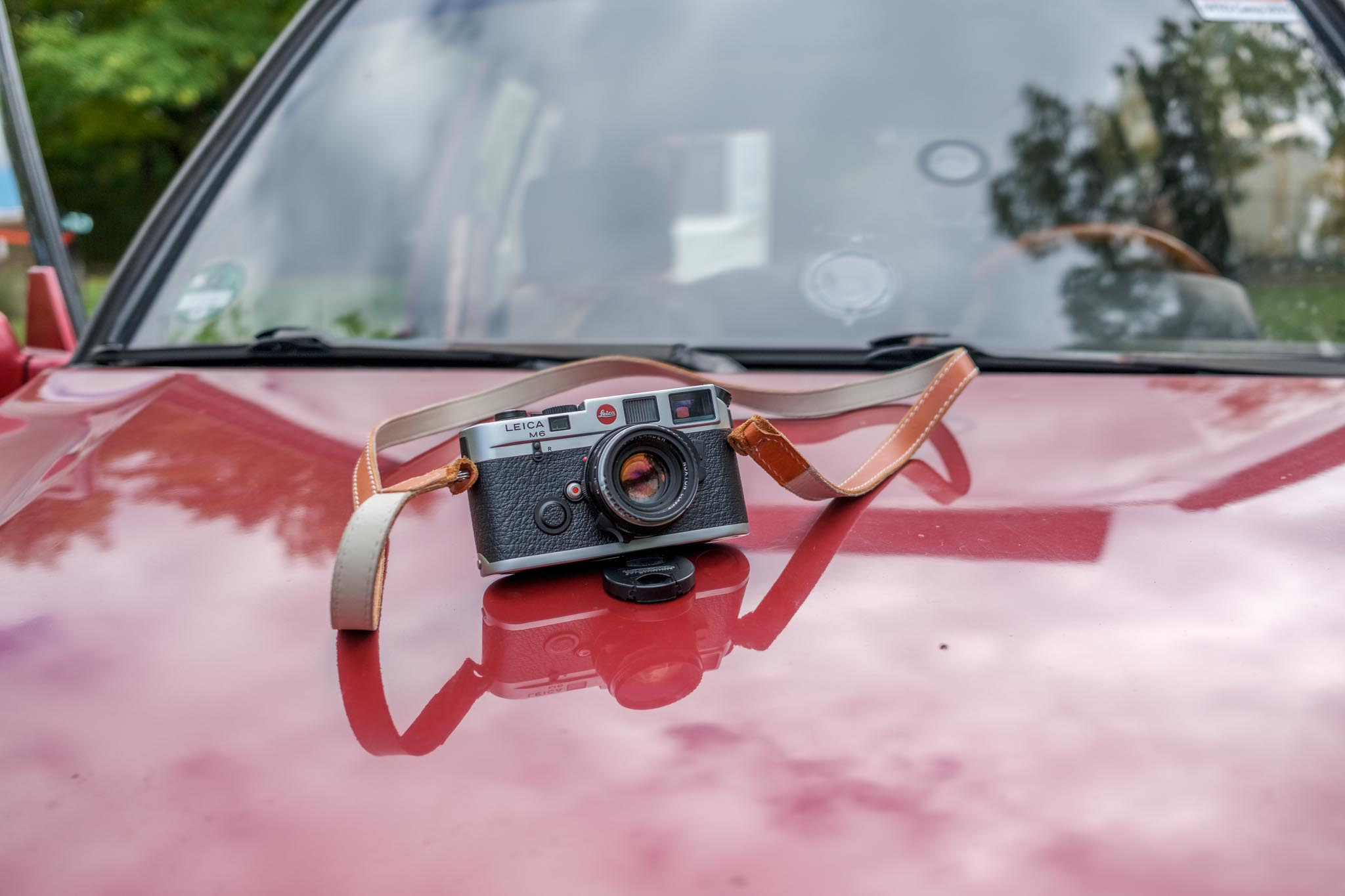 Leica M6 Kamera mit Lederriemen liegt auf der roten Motorhaube eines Autos, im Hintergrund die Windschutzscheibe mit Bäumen.