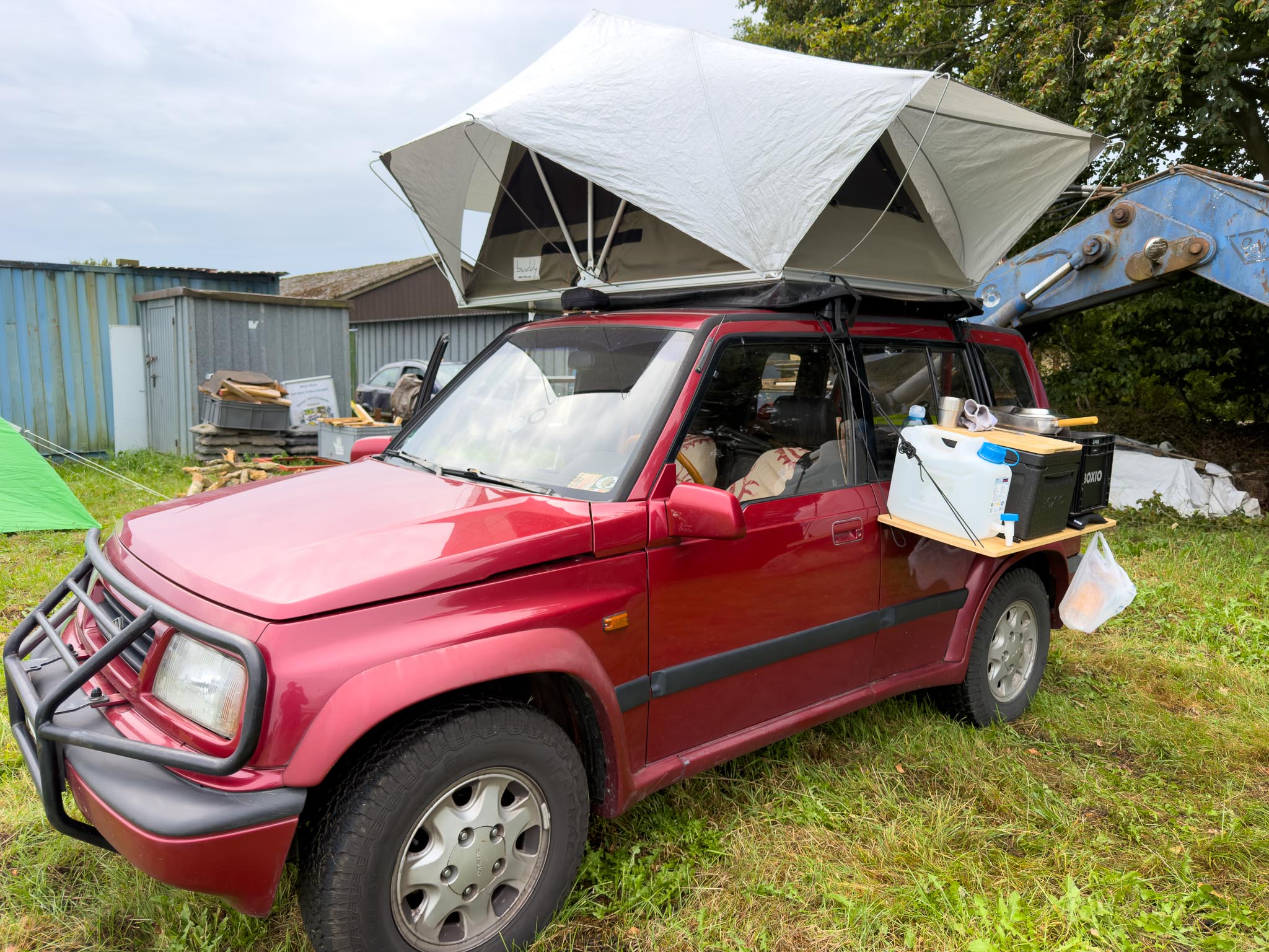 Roter Suzuki Geländewagen mit aufgeklapptem Dachzelt auf einer Wiese, seitliche Ablage mit Campingausrüstung.