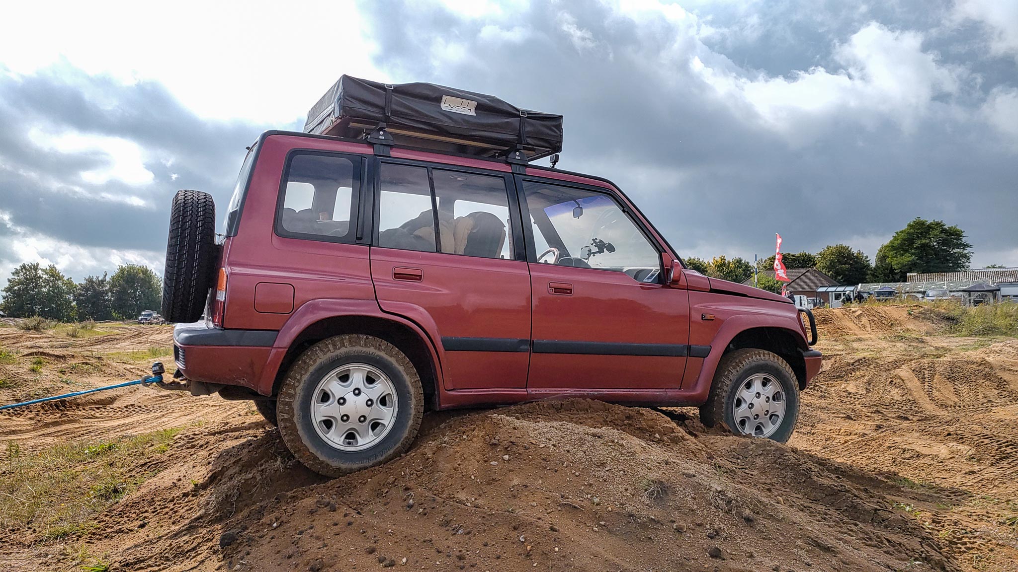 Roter Suzuki-Geländewagen mit Dachzelt überquert Sandhügel auf einem Offroad-Übungsgelände.