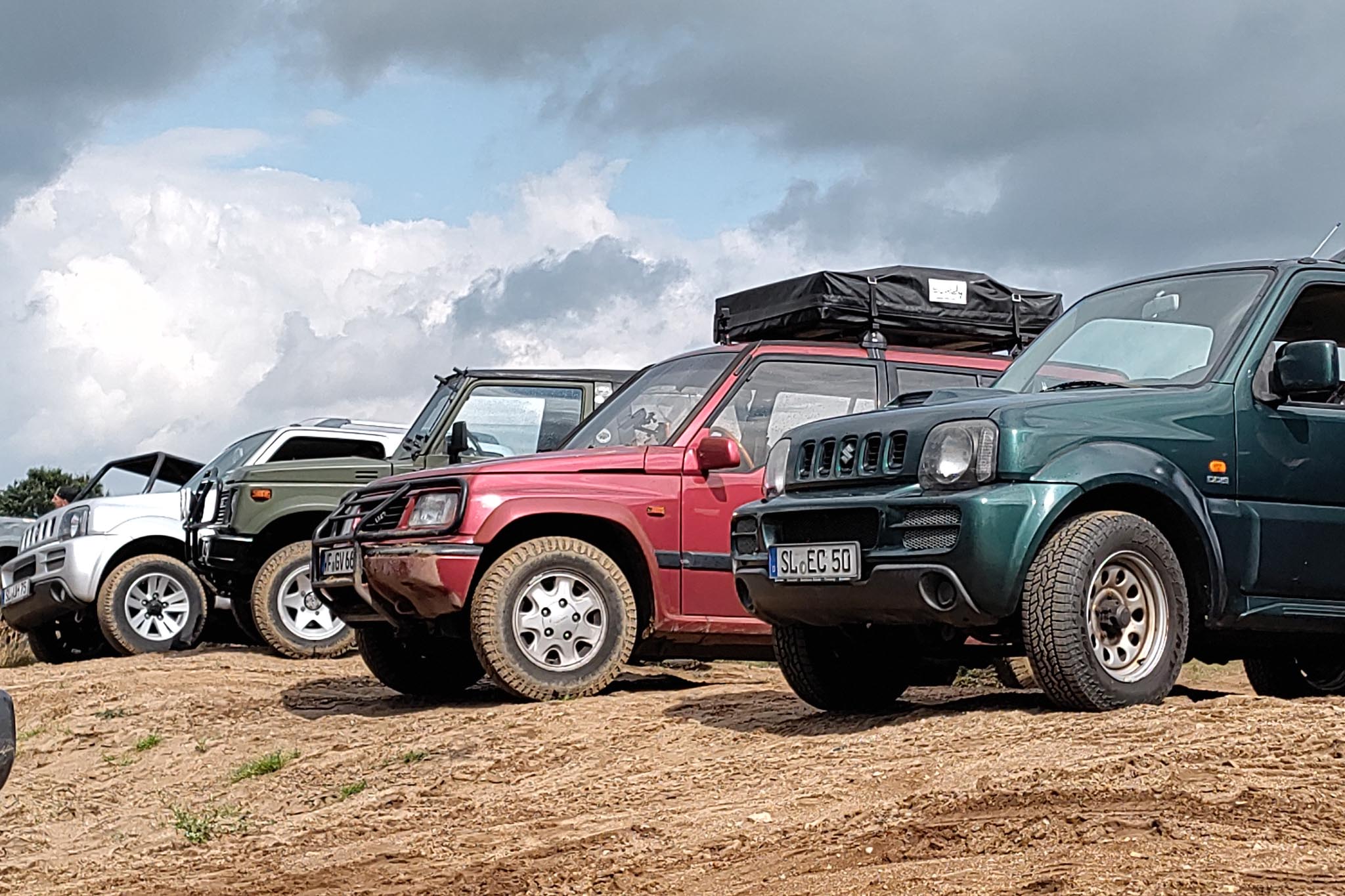 Reihe geparkter Suzuki-Geländewagen in Weiß, Oliv, Rot und Grün auf sandigem Gelände bei bewölktem Himmel.