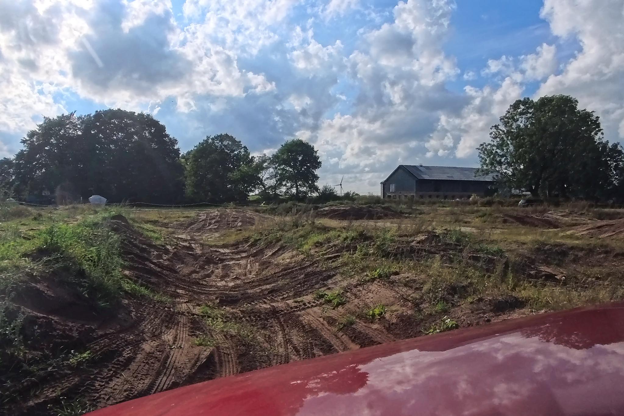 Blick über die rote Motorhaube auf ein sandiges Offroad-Geländestrecke mit tiefen Reifenspuren, im Hintergrund Bäume und eine Scheune.